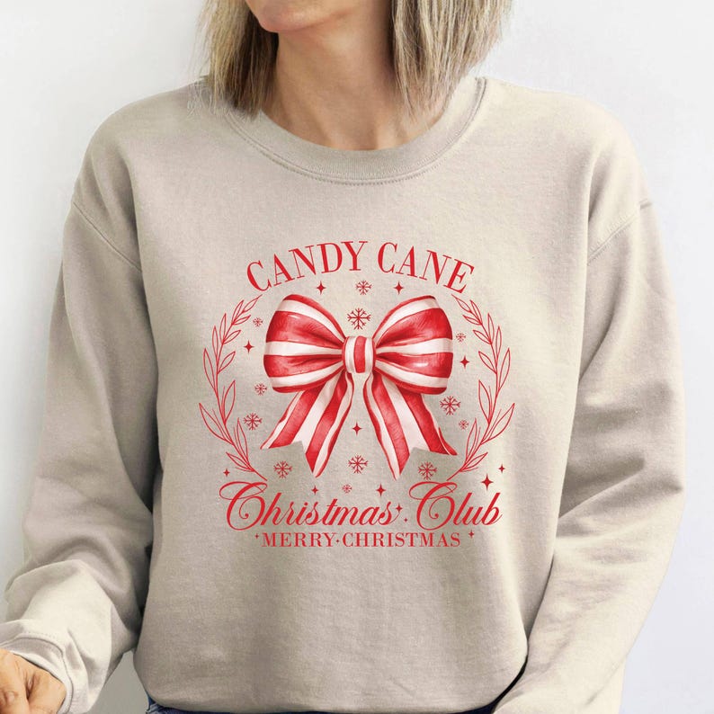 May include: A beige sweatshirt with a red and white candy cane bow design. The text "CANDY CANE Christmas Club" is printed above and below the bow, with "MERRY CHRISTMAS" at the bottom. The design is framed by a wreath.
