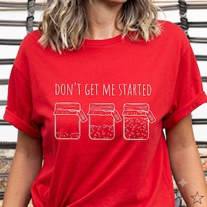 May include: A bright red short-sleeved t-shirt featuring a white graphic of three glass jars with labels. The text "DON'T GET ME STARTED" is printed in white above the jars, which show increasing levels of small dots representing contents.