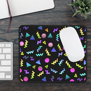 Può includere: Un tappetino per mouse nero con un motivo geometrico in colori arcobaleno. Il motivo include triangoli, zig zag e cerchi. Un mouse per computer bianco è sul tappetino per mouse.