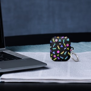 May include: A black AirPod case with a geometric pattern in pink, blue, yellow, and green. The case has a silver keychain attached.