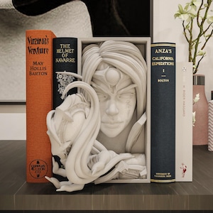 Könnte beinhalten: Ein mattweißer Buchständer mit einem skulpturalen Relief eines menschlichen Gesichts mit fließendem Haar und einer abstrakten Kreaturenform, die nach außen ragt. Der rechteckige Rahmen steht zwischen zwei gebundenen Büchern.