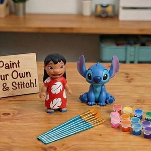 Może przedstawiać: Drewniany znak z napisem "Paint Your Own Lilo & Stitch!" jest wyświetlany obok figurek Lilo i Stitch. Widoczne są również pędzle i zestaw kolorowych farb, sugerujący zestaw rzemieślniczy.