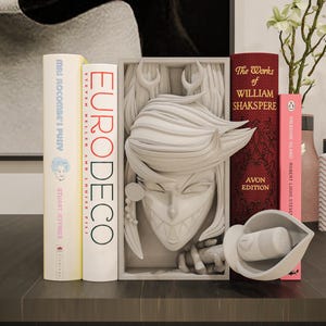 Peut inclure: Un serre-livres rectangulaire gris clair présentant un relief sculpté d'un personnage aux dents pointues, aux oreilles pointues et aux cheveux stylisés. Le serre-livres est placé entre plusieurs livres, dont Eurodeco et The Works of William Shakspeere.