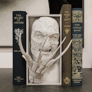 Peut inclure: Une sculpture blanche d'un personnage de film d'horreur avec de longues griffes acérées, encadrée entre plusieurs livres anciens. Les livres sont dans des tons bleu foncé et marron, avec des lettres dorées. Titres : "The Helmet of Navarre" et "The Jungle Book".