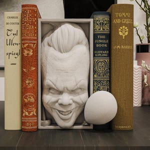 May include: A collection of books and decorative items on a dark surface. The books include titles like "Tommy and Grizel" and "The Jungle Book." A white sculpture of a smiling face is centered between the books. A white sphere is in front of the books.