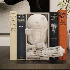May include: A white sculpture of a helmet and a blaster, displayed between several books. The books have titles like "Anza's California Expeditions" and "The Helmet of Navarre". The scene is set on a dark surface.