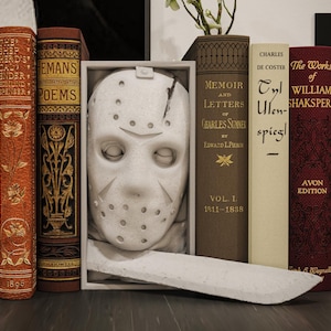 May include: A still life arrangement featuring a white hockey mask in a shadow box, a white machete, and several antique books. The books have titles like "The Works of William Shakspere" and "Memoir and Letters". The books are in various colors.