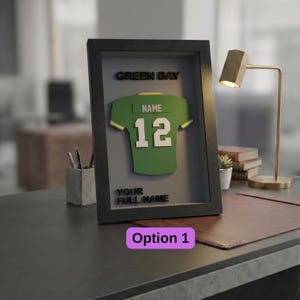 Pode incluir: Uma camisola de futebol verde e dourada emoldurada com o texto "GREEN BAY", "NAME" e "12". A moldura também inclui o texto "YOUR FULL NAME" e "Option 1". A moldura é preta e está numa secretária.