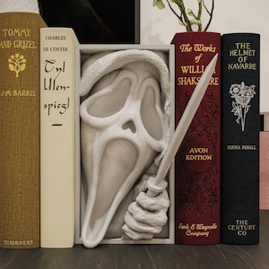 Peut inclure: Une composition décorative avec un masque de Scream blanc et un couteau argenté, entourée de livres anciens de différentes couleurs. Les livres incluent des titres comme "Tommy and Grizel" et "The Works of William Shakespeare."