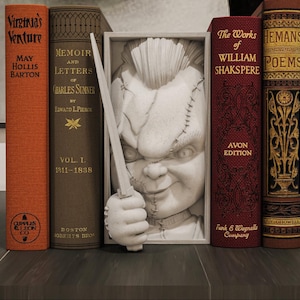 Peut inclure: Une collection de livres anciens de différentes couleurs, dont orange, marron et bordeaux. Une sculpture blanche d'une poupée tenant un couteau est placée entre les livres. Les livres ont des lettres dorées et des motifs décoratifs.