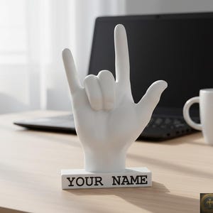 Könnte beinhalten: Eine weiße Keramik-Tasse mit schwarzem Henkel und dem Text "But First, Coffee" in fetter, schwarzer Schrift. Die Tasse steht vor einem verschwommenen Hintergrund, der eine gemütliche Umgebung suggeriert.