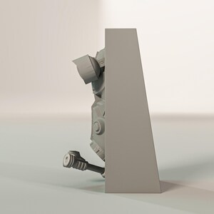 May include: A gray, three-dimensional model of a mechanical object, possibly a robot or part of a larger machine. The model has a complex design with various geometric shapes, cylinders, and circular elements. The object is partially obscured by a gray rectangular structure.