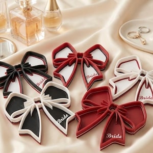 May include: Bow-shaped decorations in red, black, and white, with the word "Bride" printed on the bow's centre. The bows are arranged on a satin surface, alongside perfume bottles and a jewellery dish.