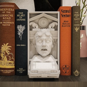 Puede incluir: Un sujetalibros decorativo con una escultura blanca de un personaje con gafas y un coche, flanqueado por libros de estilo vintage. Los libros tienen títulos como "The Riviera of the Corniche Road" y "Virginia's Venture". El sujetalibros tiene un reloj.