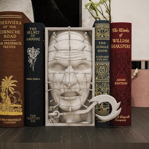 Può includere: Una collezione di libri con dorsi decorativi, tra cui titoli come "The Riviera of the Corniche Road" e "The Works of William Shakespeare". Una scultura bianca di un volto è centrata tra i libri. Un oggetto decorativo in metallo è di fronte ai libri.