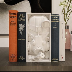 May include: A still-life arrangement featuring several books with titles like "Virginia's Venture" and "The Helmet of Navarre." A white sculpture of a gas mask is displayed between the books. Decorative vases and a framed artwork complete the scene.