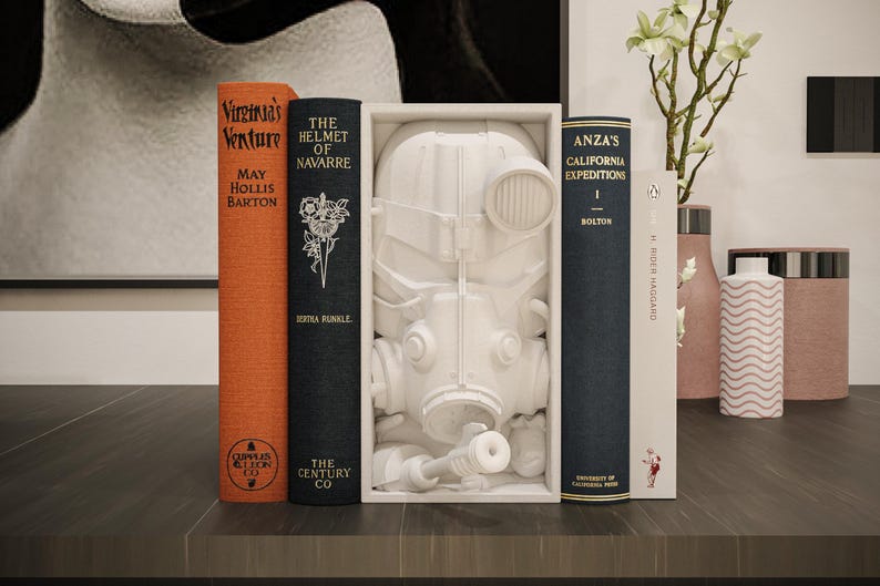May include: A still-life arrangement featuring several books with titles like "Virginia's Venture" and "The Helmet of Navarre." A white sculpture of a gas mask is displayed between the books. Decorative vases and a framed artwork complete the scene.