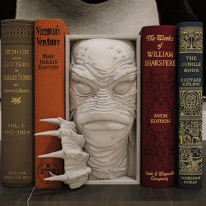 May include: A collection of vintage books with titles including "Memoir and Letters", "Virginia's Venture", "The Works of William Shakspere", and "The Jungle Book". A white creature head sculpture is centered between the books.