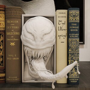 Op de afbeelding: Een decoratieve boekenplankopstelling met vintage boeken en een wit beeldhouwwerk van een wezen met een open mond en lange tong. De boeken hebben titels als "Poems" en "The Jungle Book."