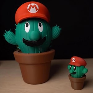 Puede incluir: Dos figuras de cactus verdes con sombreros rojos con un emblema blanco, que se asemejan a un personaje popular de videojuegos. El cactus más grande está en una maceta marrón, y una versión más pequeña está al lado, también en una maceta marrón.