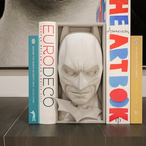 May include: A gray Batman head bookend with the Batman logo, flanked by books. The books include "Eurodeco", "Around the World in Eighty Days", "The Heart Book", and "The Middle of the Earth". The bookends are on a dark surface.