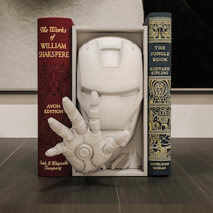 May include: A white Iron Man helmet and hand sculpture displayed between two vintage books. The books are titled "The Works of William Shakspere" and "The Jungle Book" with gold lettering. The sculpture is in a white frame.