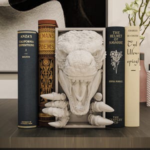 May include: A white bookend featuring a sculpted dinosaur head with an open mouth and sharp claws extending outward. The dinosaur figure is mounted within a rectangular frame, positioned between several hardcover books on a flat surface.