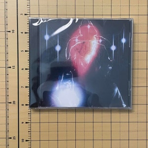 May include: A CD case with a dark, abstract design. The artwork features a central red and white shape with radiating lines and glowing orbs against a black background. The CD is sealed in plastic.