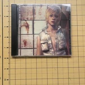 May include: A CD case featuring the cover art for the "Silent Hill 3 Original Soundtracks" album. The artwork depicts a woman with short blonde hair wearing a sleeveless shirt. The background shows a rusty, industrial setting.