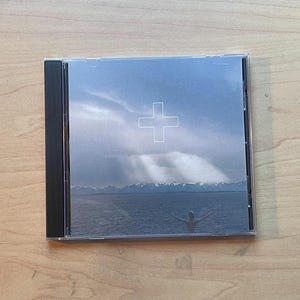 May include: A CD case featuring a serene landscape with a cloudy sky and a white cross symbol. The image depicts a person with outstretched arms in front of a body of water and mountains. The CD case is clear with a black spine.