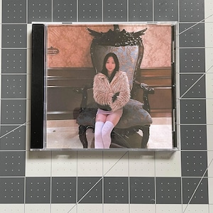 May include: A CD case featuring a photograph of a person wearing a pink fur coat, seated on an ornate chair. The background shows a patterned wall. The CD case is black on the spine.