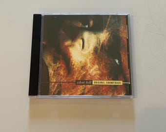May include: A compact disc jewel case featuring the Silent Hill Original Soundtrack. The cover art displays a dark, textured, and abstract image in shades of brown, black, and gold. The text Silent Hill Original Soundtrack is printed in black.