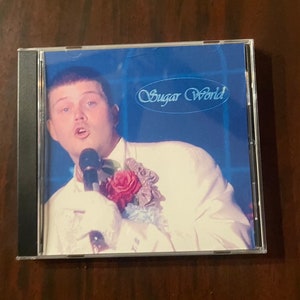 May include: A CD case with a blue background and a man in a white suit holding a microphone. The text "Sugar World" is written in a cursive font on the case.
