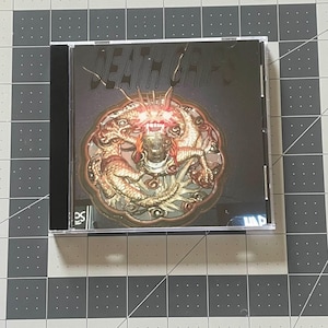 May include: A CD case featuring the band name "DEATH GRIPS" in white text on a dark background. The album art showcases a detailed, ornate design with a central figure and intricate details in shades of gold and red.