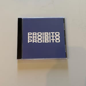 May include: A square CD jewel case with a clear plastic front and a black spine. The insert features a solid periwinkle blue background with the word PROIBITO printed in white capital letters in three stacked rows across the center.