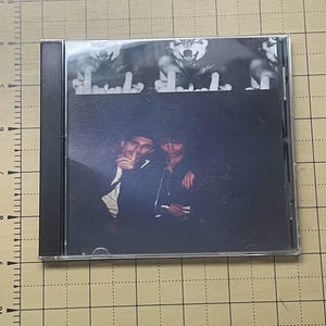 May include: A CD case featuring a black and white image. The top portion shows a repeating image of a wolf with its middle finger raised. The bottom portion depicts two people in dark clothing.