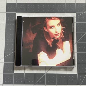 May include: A CD case featuring a woman with red lipstick and a black top. The image is in a clear plastic case with a black spine. The background is a dark, blurred setting.