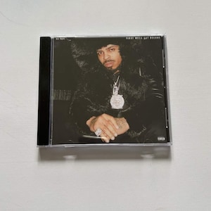 May include: A CD case featuring a portrait of a person wearing a fur coat and jewelry. The text "RX PAPI" is visible on the left, and "FIRST WEEK OUT DELUXE" is on the top right. The CD case is black.