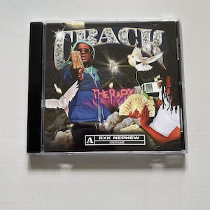 May include: A CD case featuring the album cover for "TRAC" by RXK Nephew. The cover art depicts a person holding money, with the word "THERAPY" below. The album title is in a stylized font with dollar bill accents.