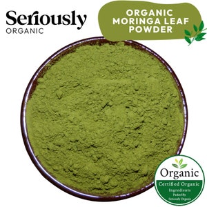 May include: A close-up of a brown bowl filled with bright green organic moringa leaf powder. The powder is finely ground and has a smooth texture. The bowl is sitting on a white background. The text "ORGANIC MORINGA LEAF POWDER" is written in white letters on a green background above the bowl. The text "Seriously Organic" is written in black letters on a white background in the top left corner of the image.