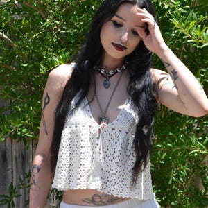 May include: A white crochet top with a plunging neckline and a matching white skirt. The top has a delicate, open weave design. The person is wearing multiple necklaces and has tattoos on their arms. The background features green foliage and a wooden fence.
