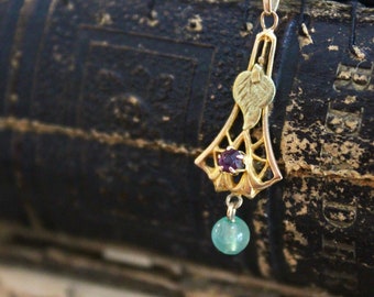 May include: A gold pendant with a green gemstone and a red gemstone. The pendant is hanging from a chain and is resting on a worn, black book.