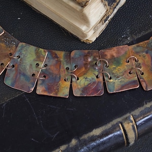 May include: A handcrafted necklace featuring square copper links with a colorful, iridescent finish. The links are connected by small metal rings. The necklace is displayed on a dark surface with a vintage book in the background.
