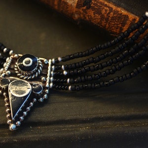 May include: A close-up of a black beaded necklace with a silver pendant. The pendant has a geometric design with black and silver accents. The necklace has multiple strands of small black beads.