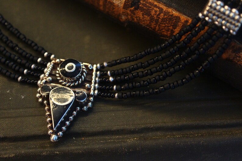 May include: A close-up of a black beaded necklace with a silver pendant. The pendant has a geometric design with black and silver accents. The necklace has multiple strands of small black beads.