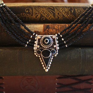 May include: A multi-strand black beaded necklace with a silver-toned pendant. The pendant features a central black stone, a circular design, and a triangular bottom. The necklace is displayed on a stack of antique books.