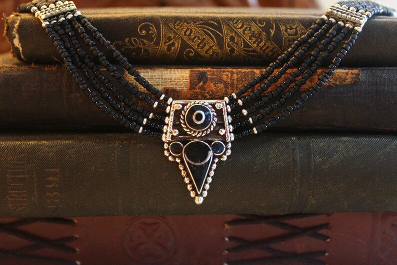 May include: A multi-strand black beaded necklace with a silver-toned pendant. The pendant features a central black stone, a circular design, and a triangular bottom. The necklace is displayed on a stack of antique books.