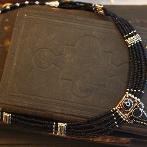 May include: A black beaded necklace with silver-toned accents. The necklace features multiple strands of small black beads and a geometric pendant with black and silver details. The necklace is displayed on a dark, textured surface.