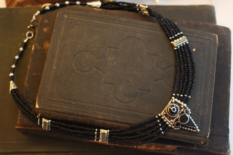 May include: A black beaded necklace with silver-toned accents. The necklace features multiple strands of small black beads and a geometric pendant with black and silver details. The necklace is displayed on a dark, textured surface.