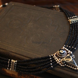 May include: A multi-strand necklace with black beads and silver-toned accents. The pendant features a geometric design with black and silver elements. The necklace is displayed on a dark, textured surface.